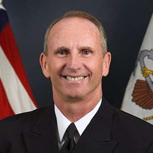 Portrait of a smiling middle-aged man in a naval uniform with American and Navy flags in the background.