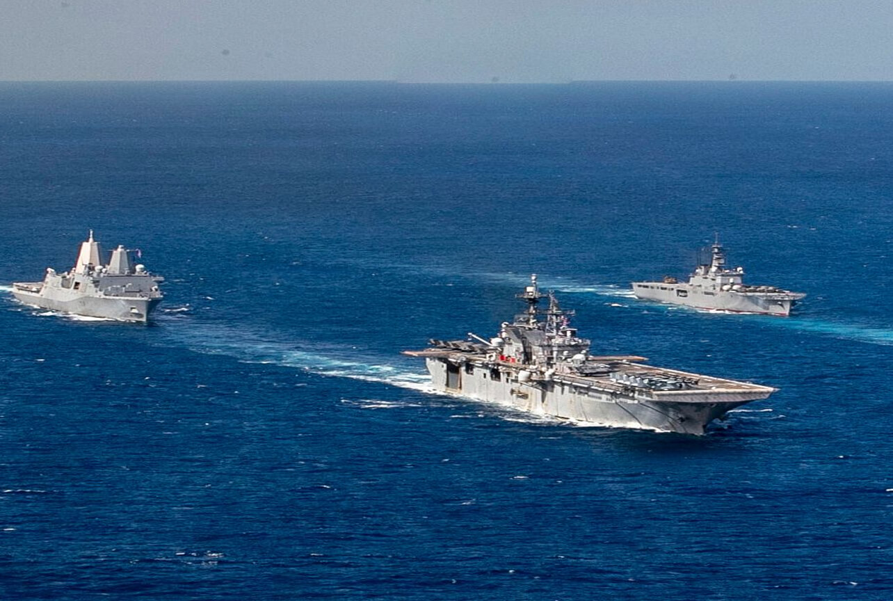 Three naval warships sailing in formation on the open sea, with the largest aircraft carrier in the center and two smaller ships flanking it on each side.