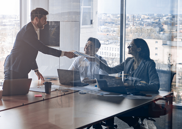 TQI team in a meeting room with laptops, one handing a document to a seated colleague, with a city view through large windows.