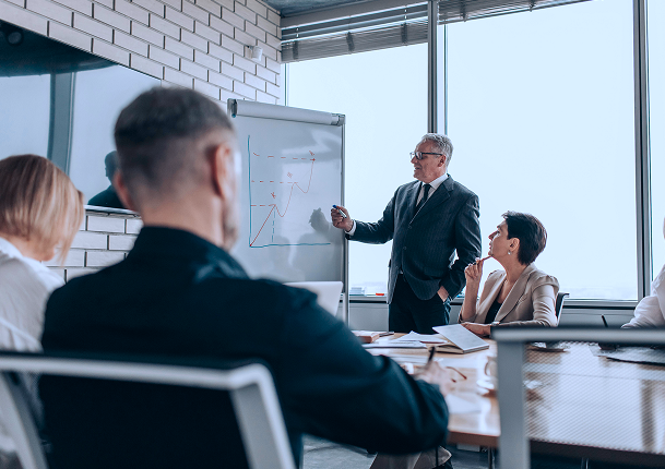 TQI team presenting a growth chart on a flip chart to colleagues seated around a conference table in a modern office with large windows.