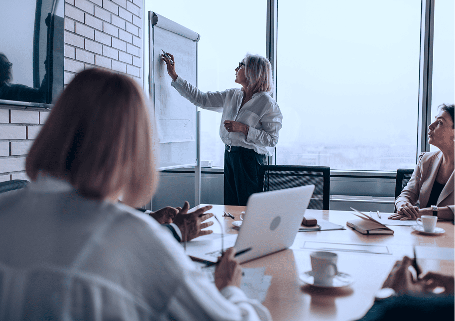 A businesswoman standing and writing on a flip chart in a conference room while colleagues sit around a table with laptops, notebooks, and coffee cups, attentively listening.