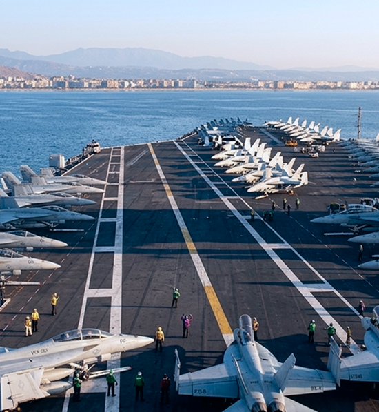 View of an aircraft carrier deck with multiple fighter jets lined up on both sides and crew members in colored uniforms standing and walking around, the sea and distant city skyline visible in the background.