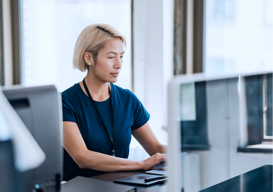Person with short blond hair wearing a dark blue shirt and a black lanyard, working on a laptop in a bright office space with large windows.