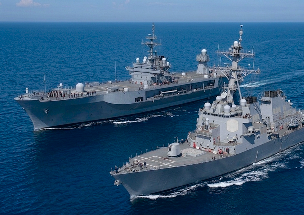 Two gray naval warships sailing side by side on a calm blue ocean under a clear sky, with personnel visible on the decks.