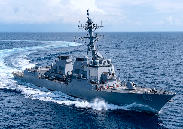 Gray naval warship numbered 68 moving swiftly through the ocean water with visible waves and blue sky with clouds.