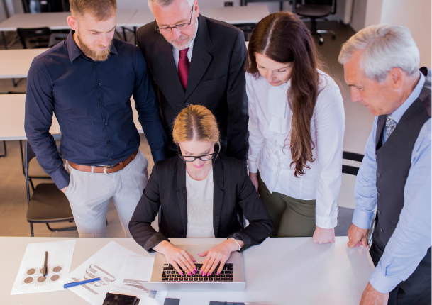 A woman in glasses types on a laptop at a table while four colleagues stand around her, watching attentively in a professional office setting.