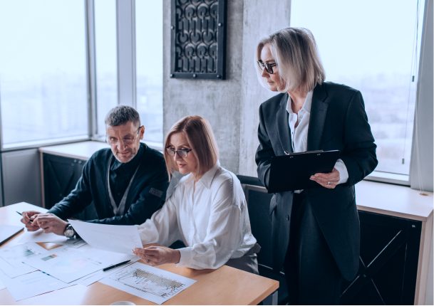 Three professionals in an office reviewing charts and documents; one woman seated holding a paper, a man seated next to her, and another woman standing with a clipboard, all focused on the work.