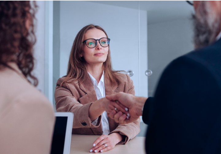 A woman in glasses and a brown blazer is shaking hands with a man across a table in an office setting, with another person partially visible in the foreground.