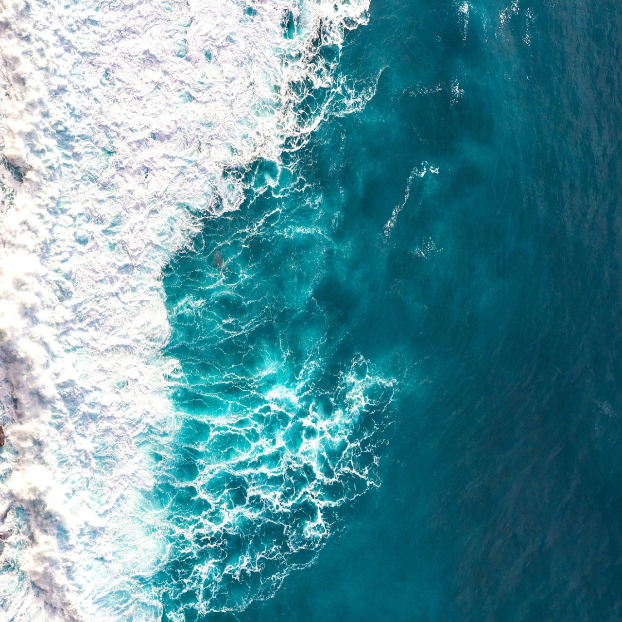 Aerial view of ocean waves crashing and creating white foam on turquoise blue water.