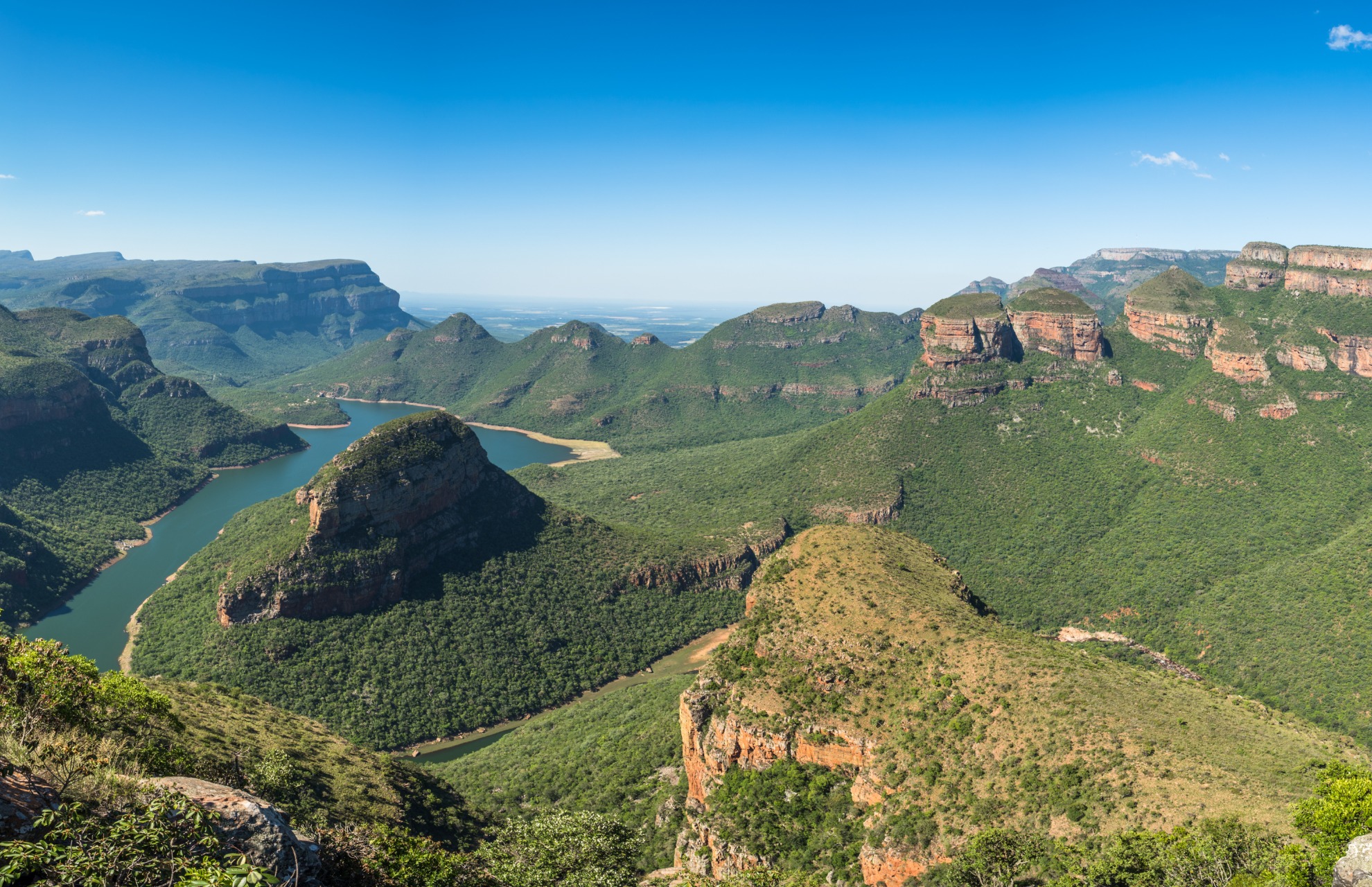 Panoramic view of a lush green canyon with a winding river flowing between steep rocky cliffs under a clear blue sky.