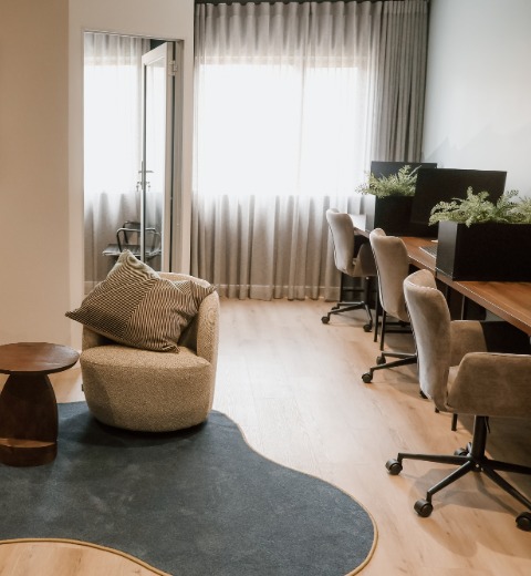 Modern office space with light wood flooring, a soft textured armchair with a striped pillow, a small wooden side table, a blue rug, a row of gray swivel chairs at a long wooden desk, computer monitors, and green plants in black planters under a large window with sheer curtains.
