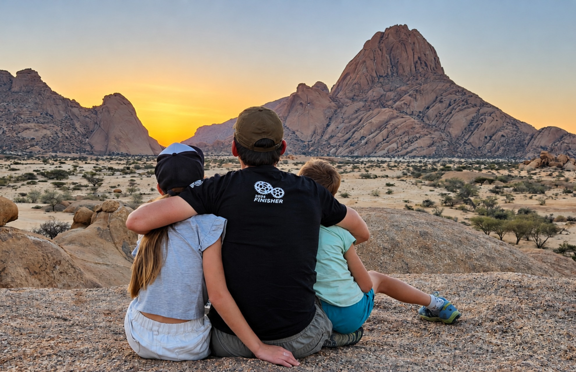 Three people sitting on a rocky surface facing large rock formations at sunset in a desert landscape, with the person in the middle wearing a black shirt and olive cap embracing the other two.