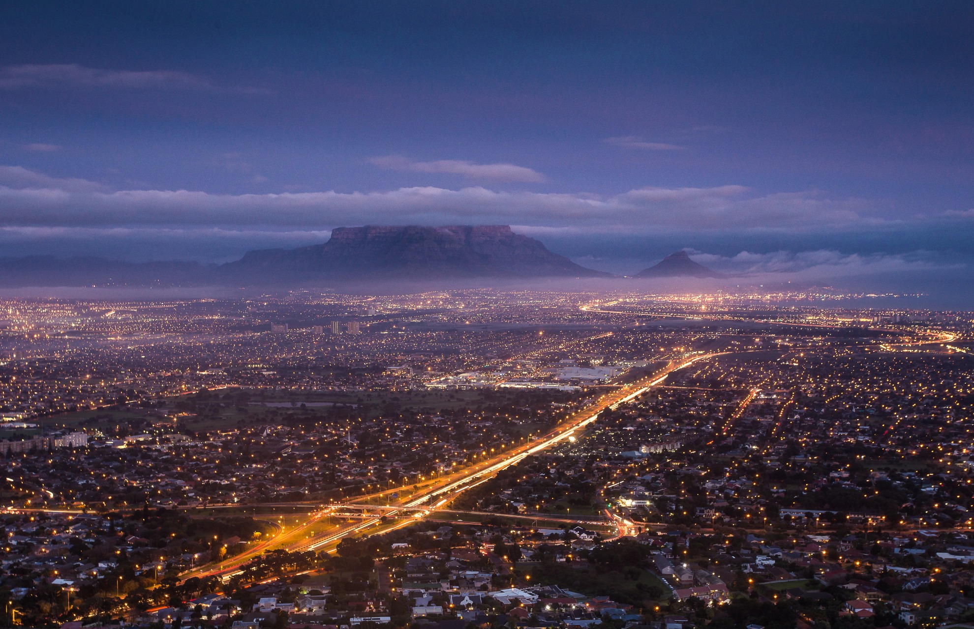 A wide aerial night view of a city with illuminated streets and buildings, featuring a brightly lit highway running through the city toward a misty mountain range in the background under a dark blue sky.
