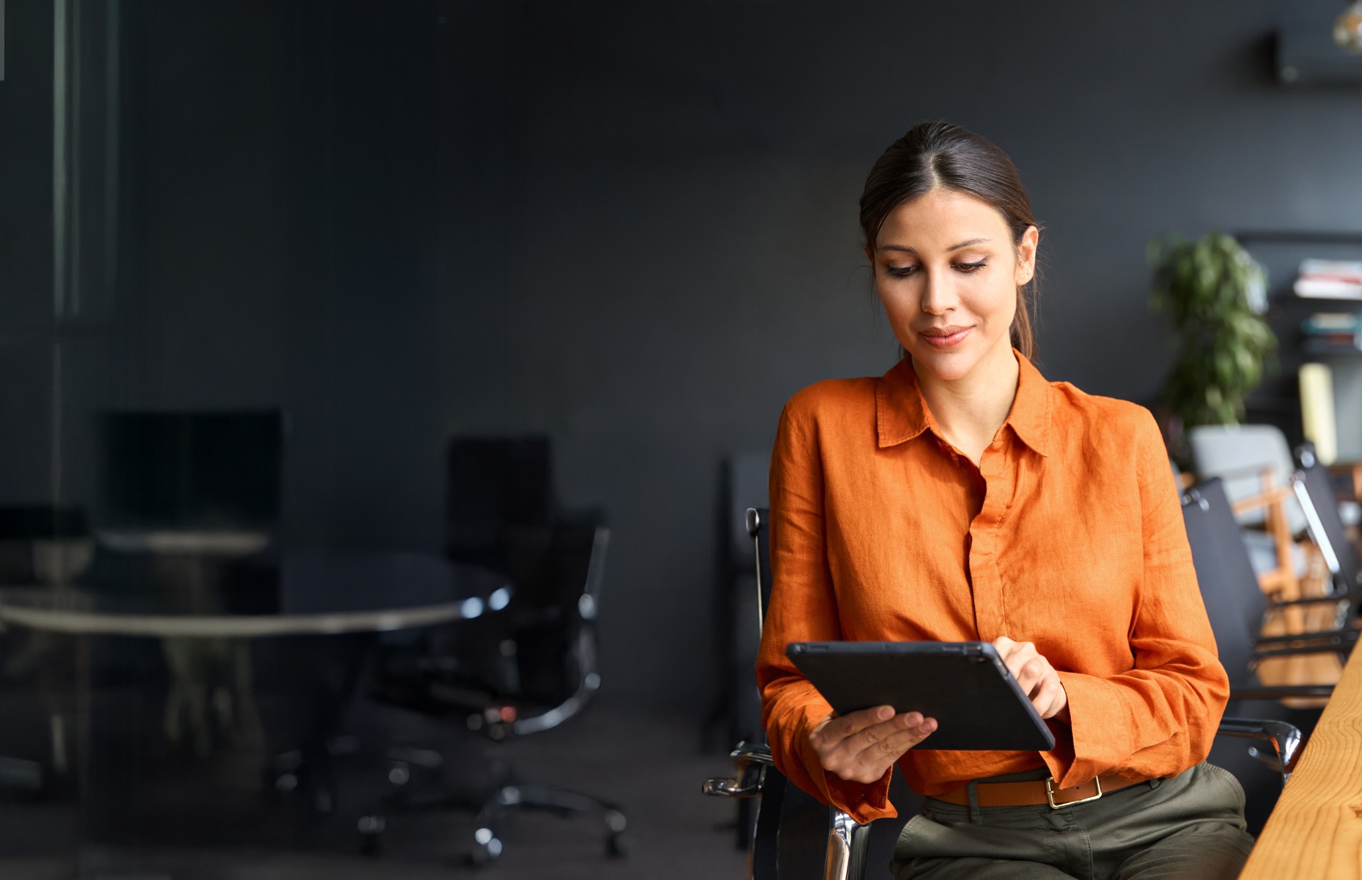 A woman with dark hair tied back wearing an orange blouse and olive pants sits in an office chair at a wooden desk, looking at and using a tablet with a slight smile, with blurred office chairs and a plant in the background under soft lighting.