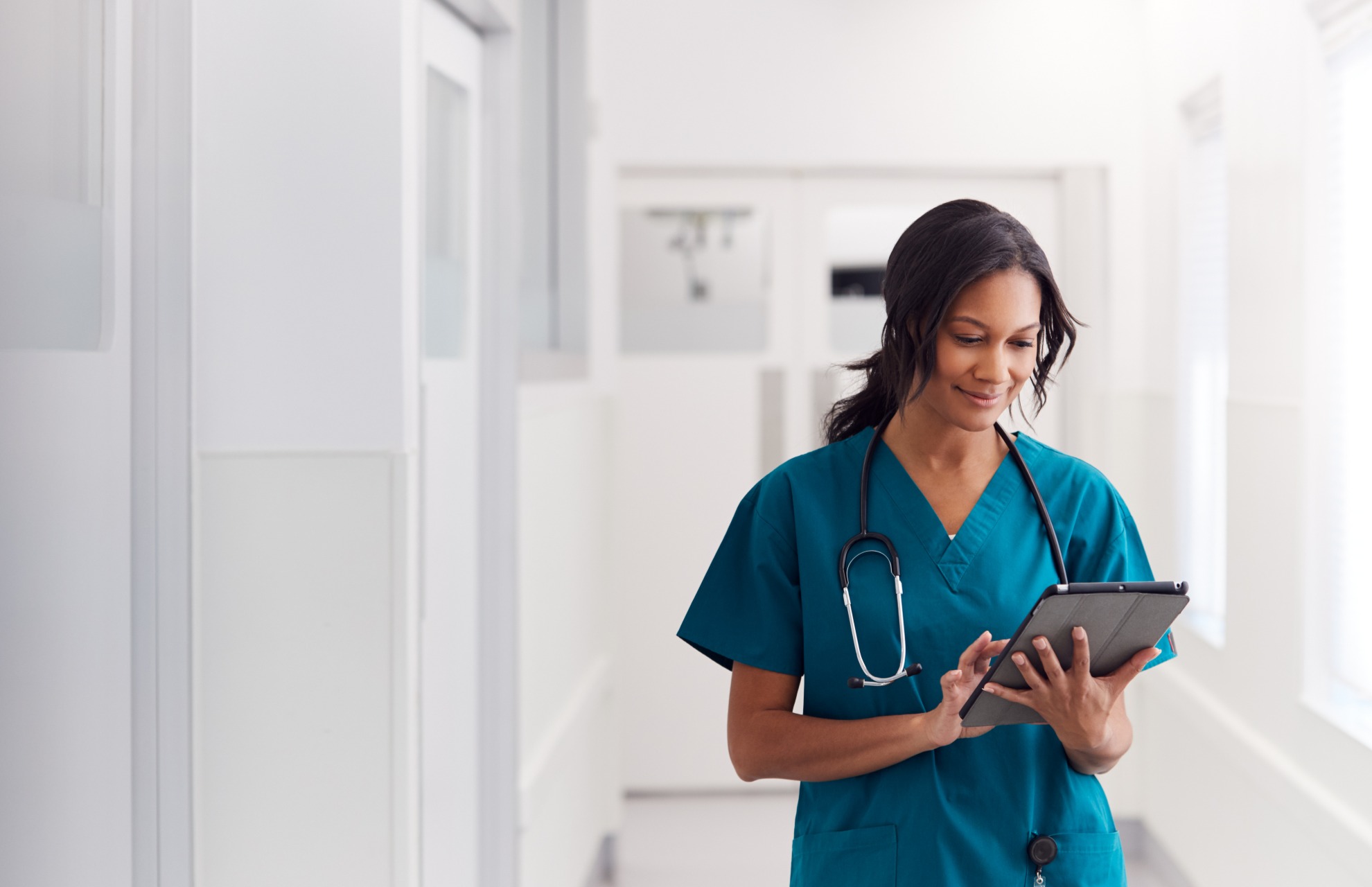 Female medical professional in teal scrubs with a stethoscope around her neck, standing in a bright hospital hallway and using a digital tablet.