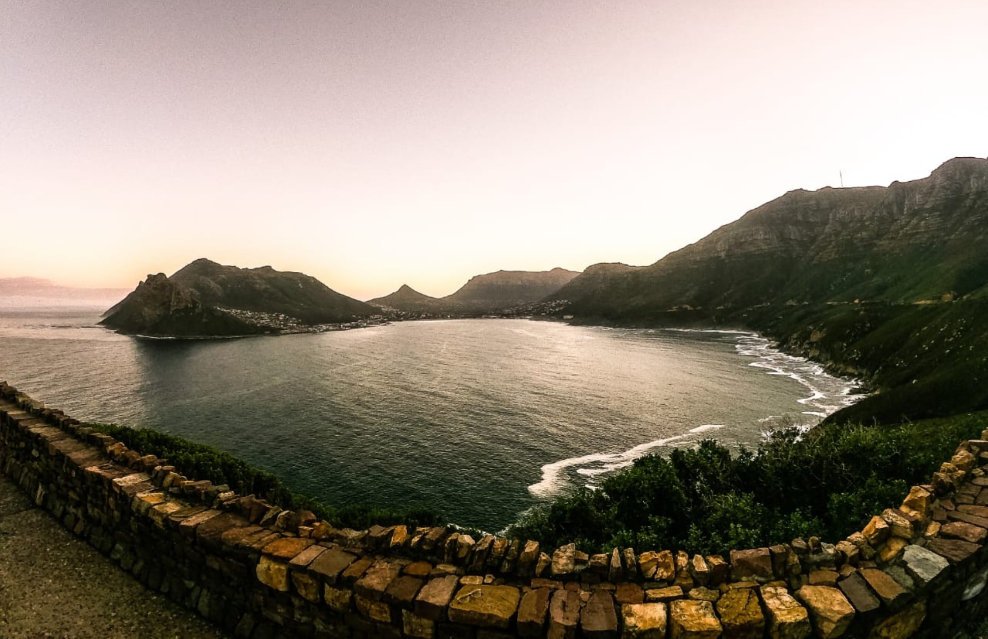 Wide view of a calm bay surrounded by green mountains under a clear sky at sunset, with a stone wall in the foreground.