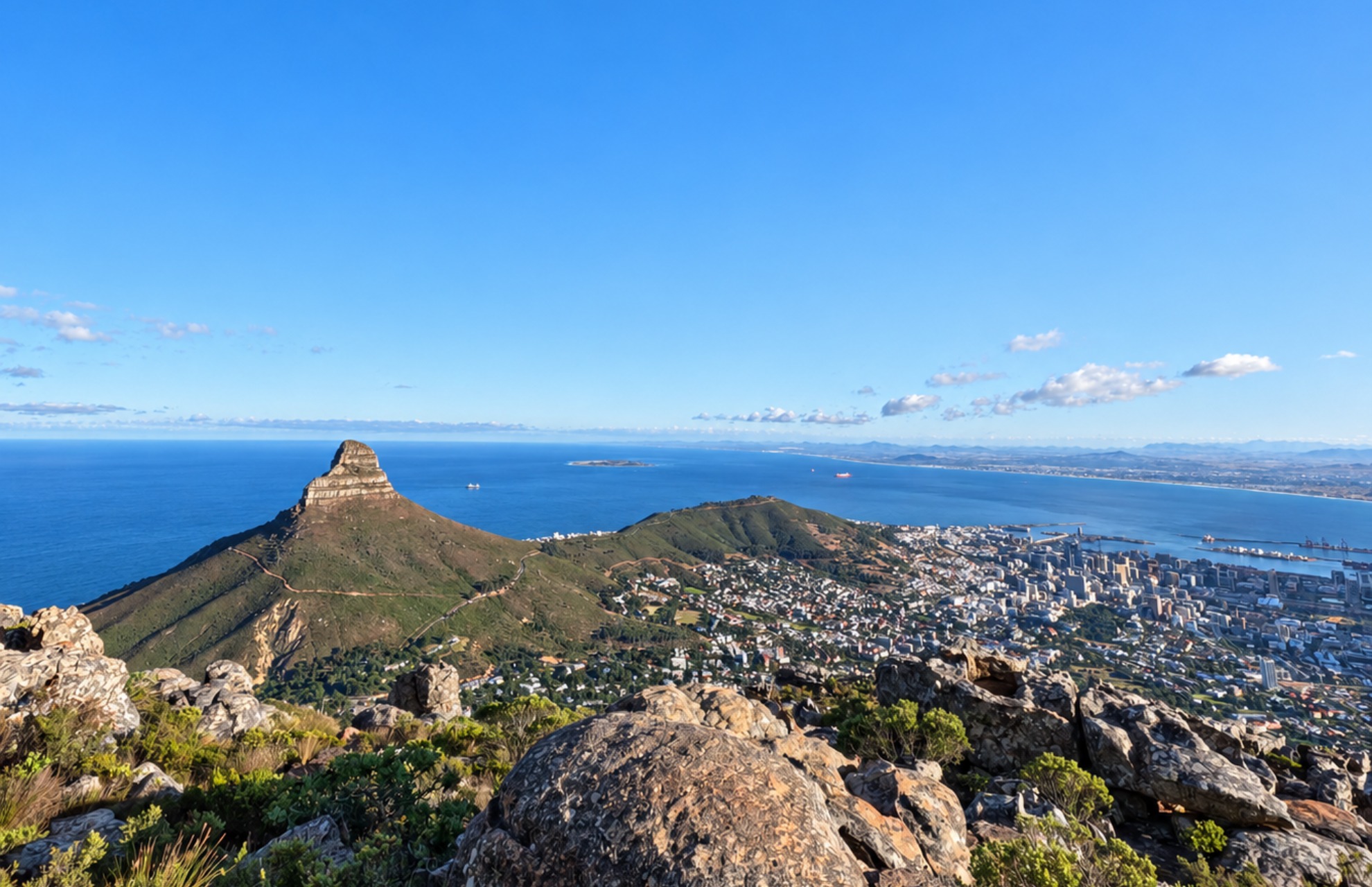 View from a rocky mountain overlooking a green hill with a distinct peak, a sprawling city with buildings, and a large body of water under a clear blue sky with some scattered clouds.