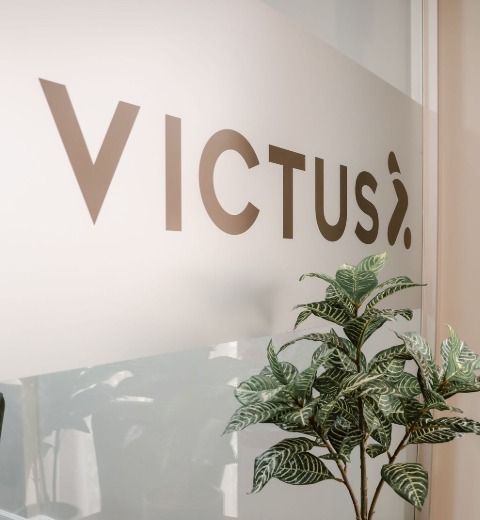 Frosted glass panel with the word 'VICTUS' in large letters, partially obscured by a leafy green plant in front.