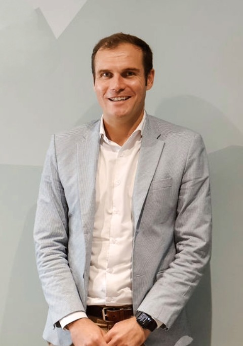 Smiling man with short brown hair wearing a light gray blazer over a white shirt, standing against a plain light gray wall.