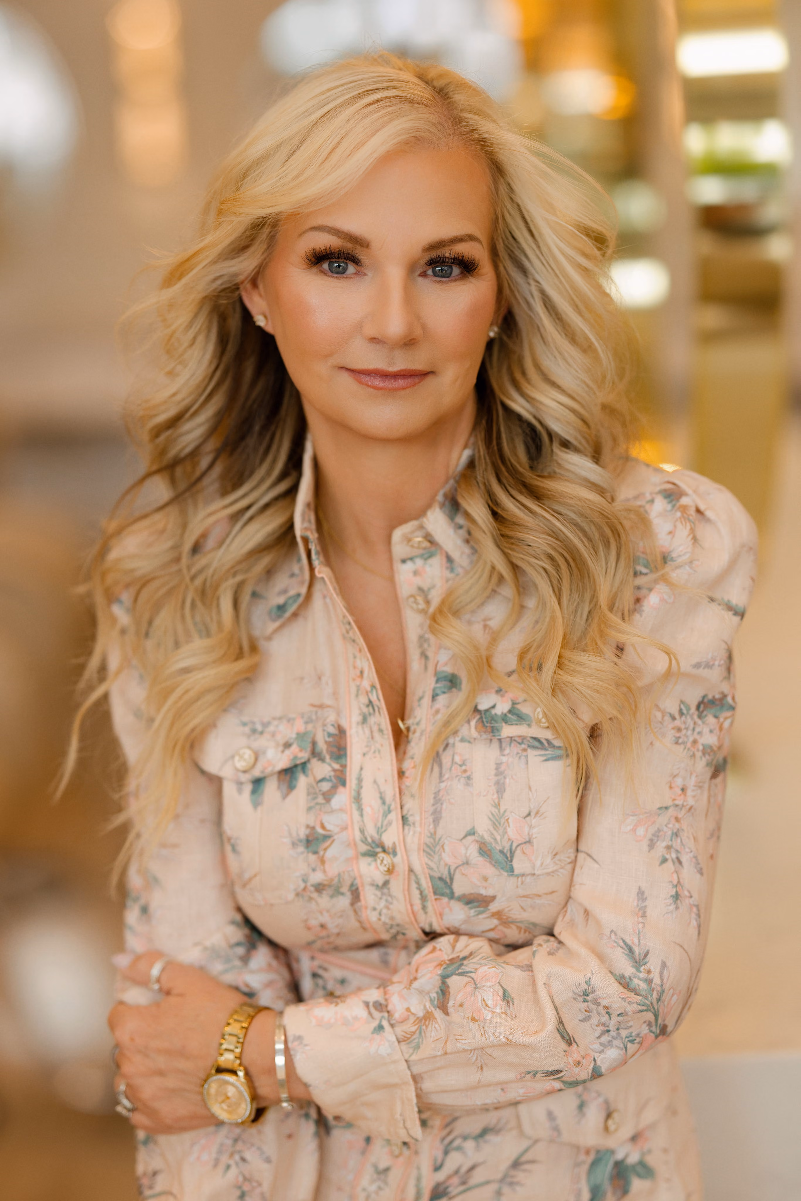 Blonde woman with curled hair wearing a floral blouse, gold watch, and light makeup in a soft-lit setting.