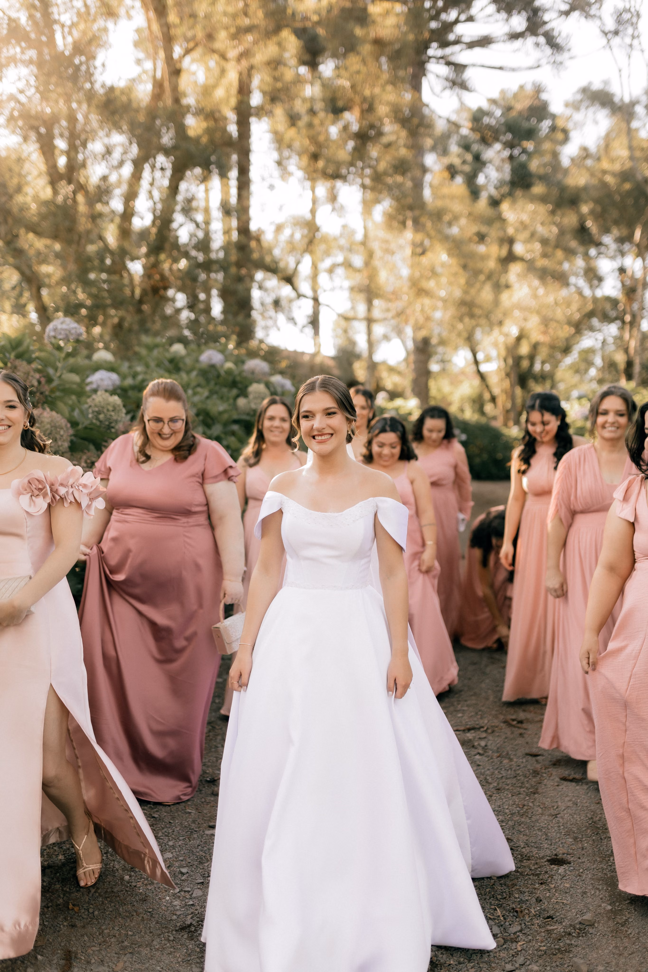 Bride in white dress smiling, surrounded by bridesmaids wearing various shades of pink dresses outdoors.