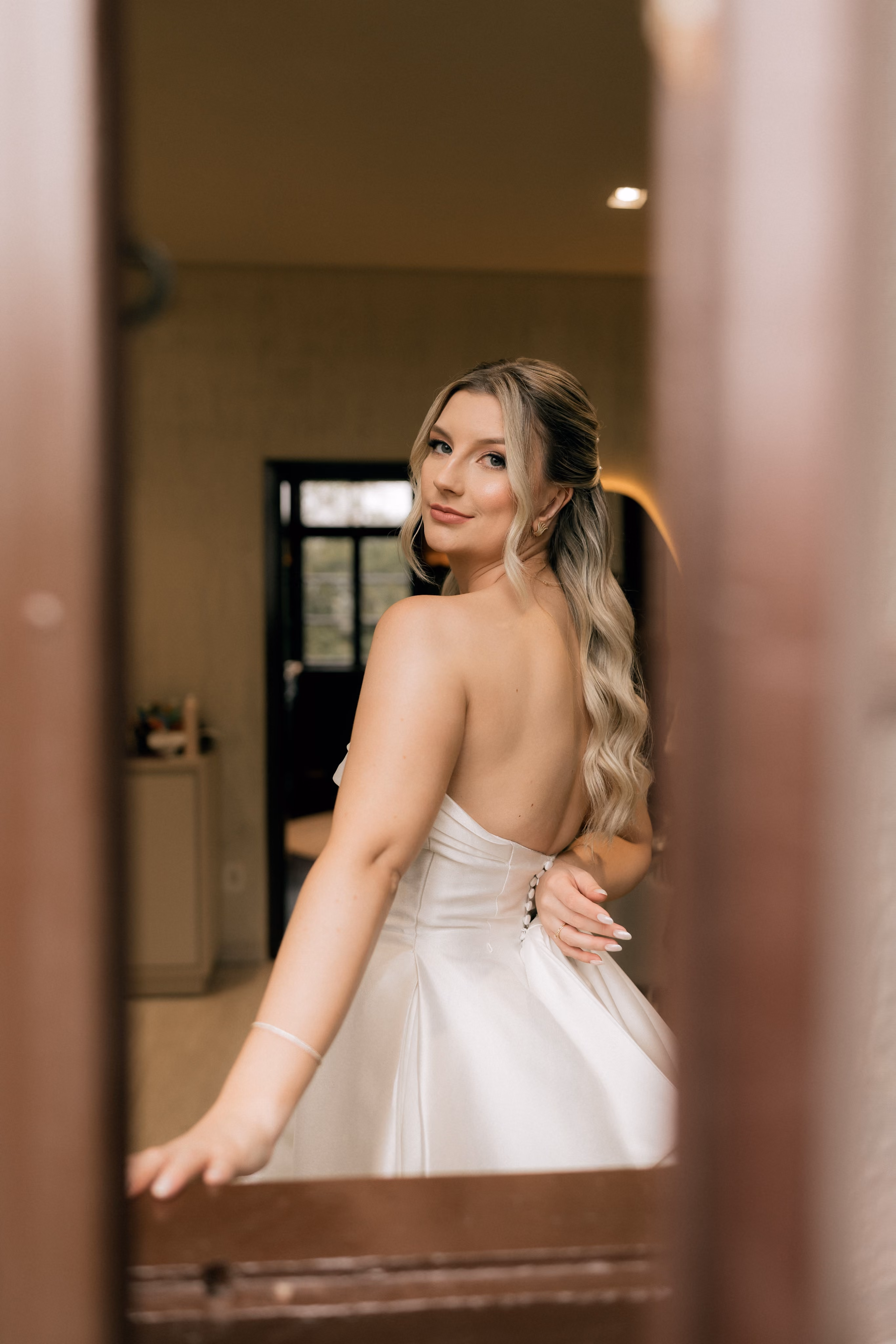 Bride in a strapless white gown looks over her shoulder inside a softly lit room.