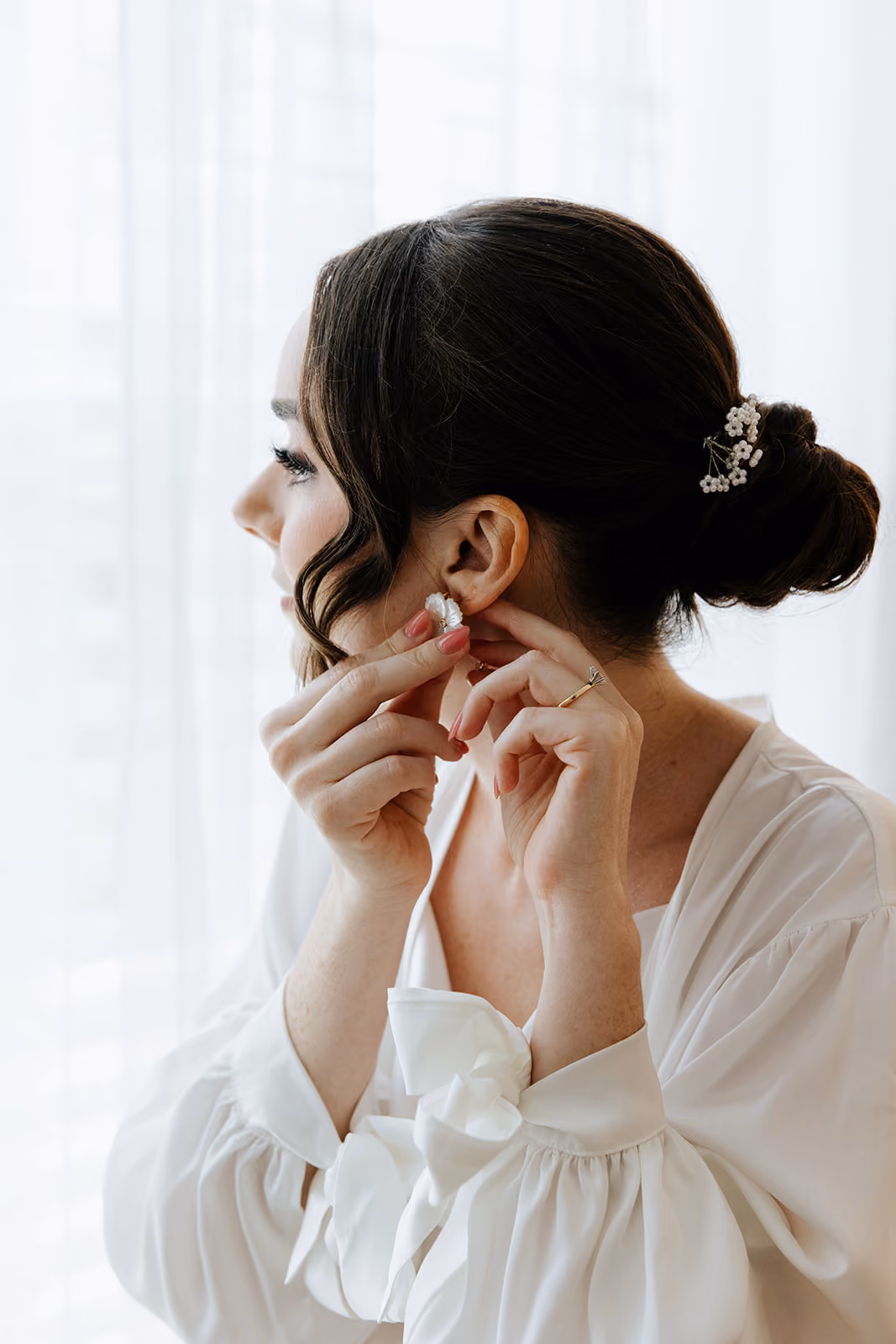Bride in a white robe fastening an earring, her dark hair in a low pinned bun