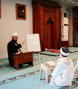 Muslim man teaching Quran in a mosque to a boy wearing traditional white clothing and cap.