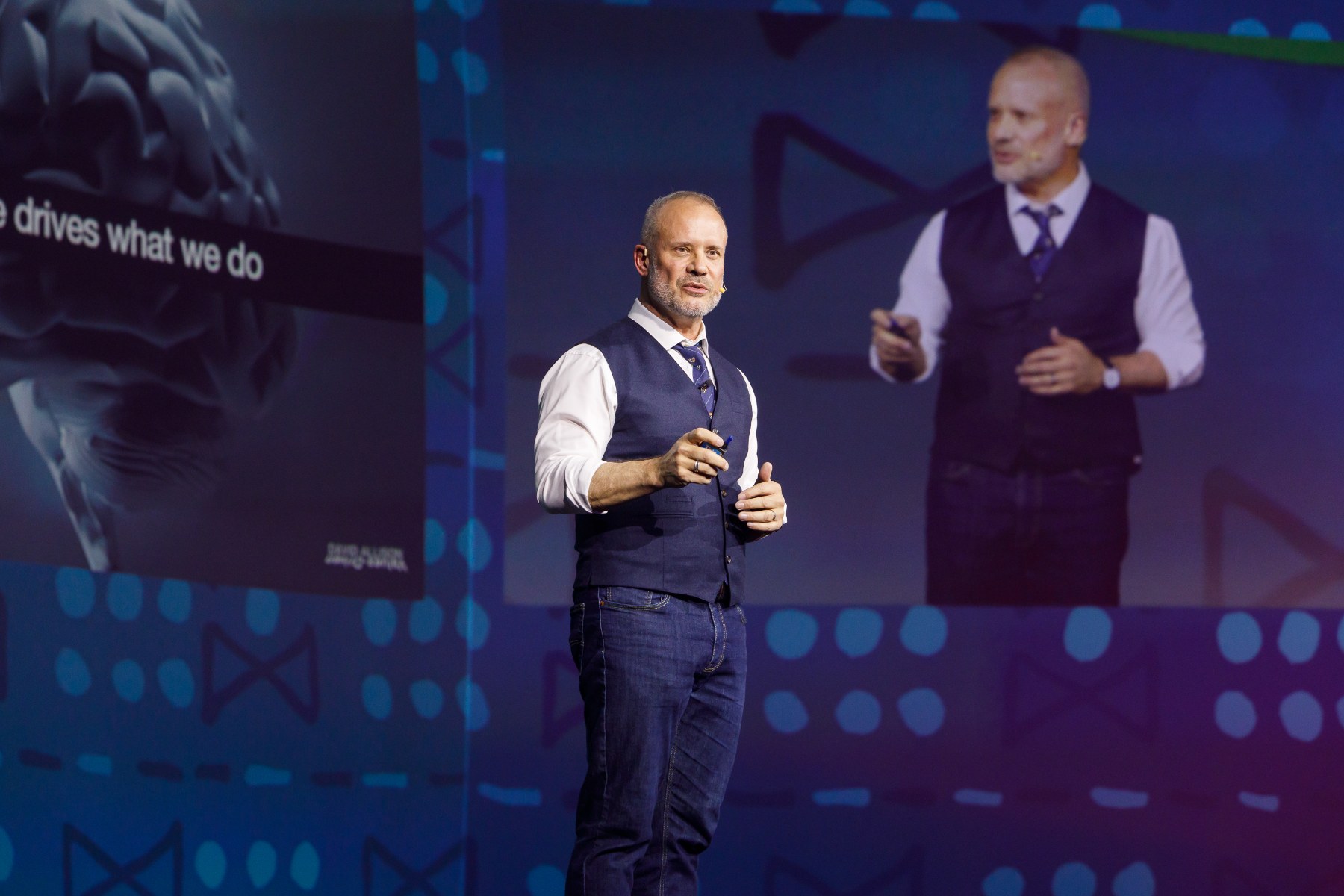 David Allison speaking on stage, shown on the big screen behind him