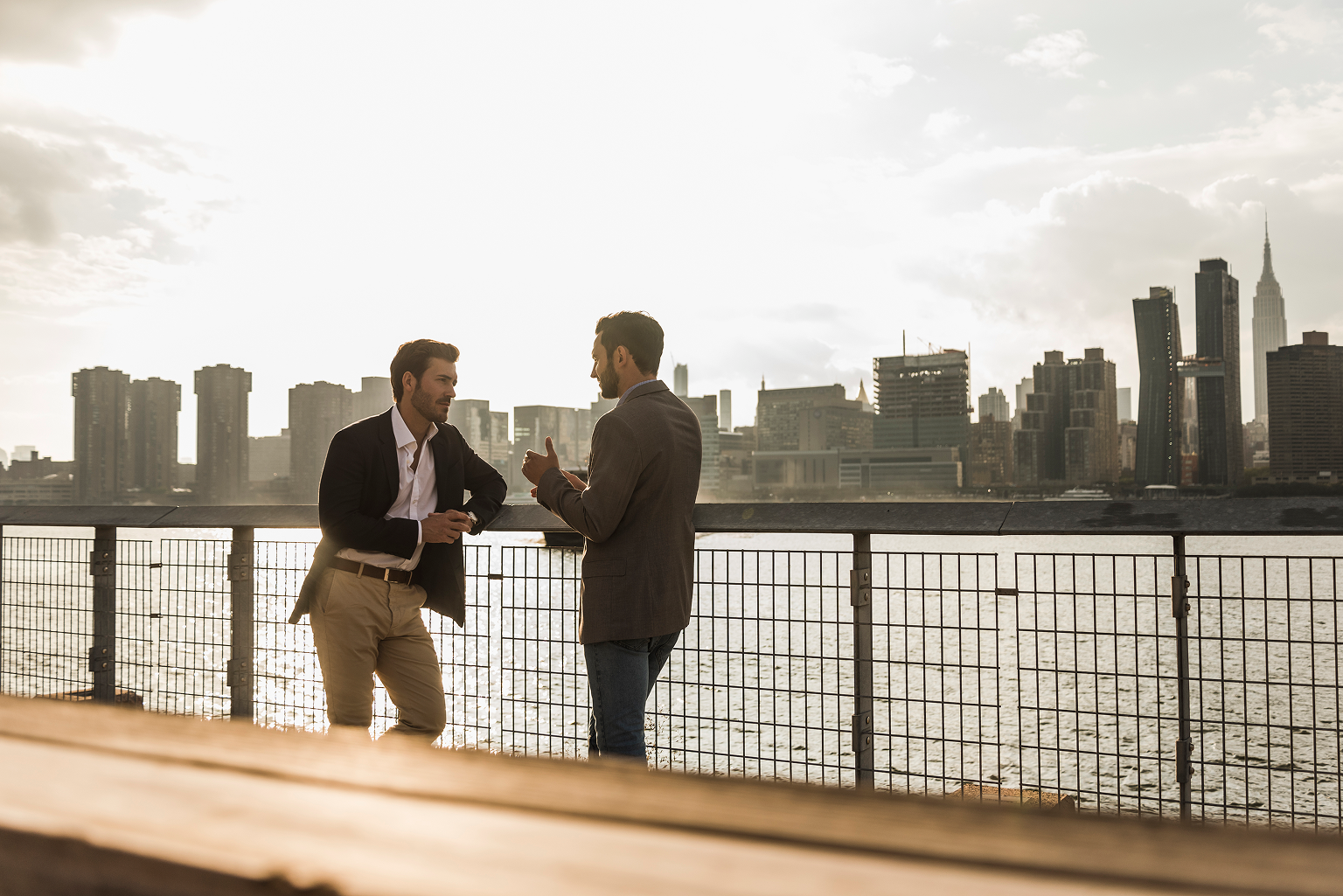 Two men in conversation at a waterfront railing with the Manhattan skyline