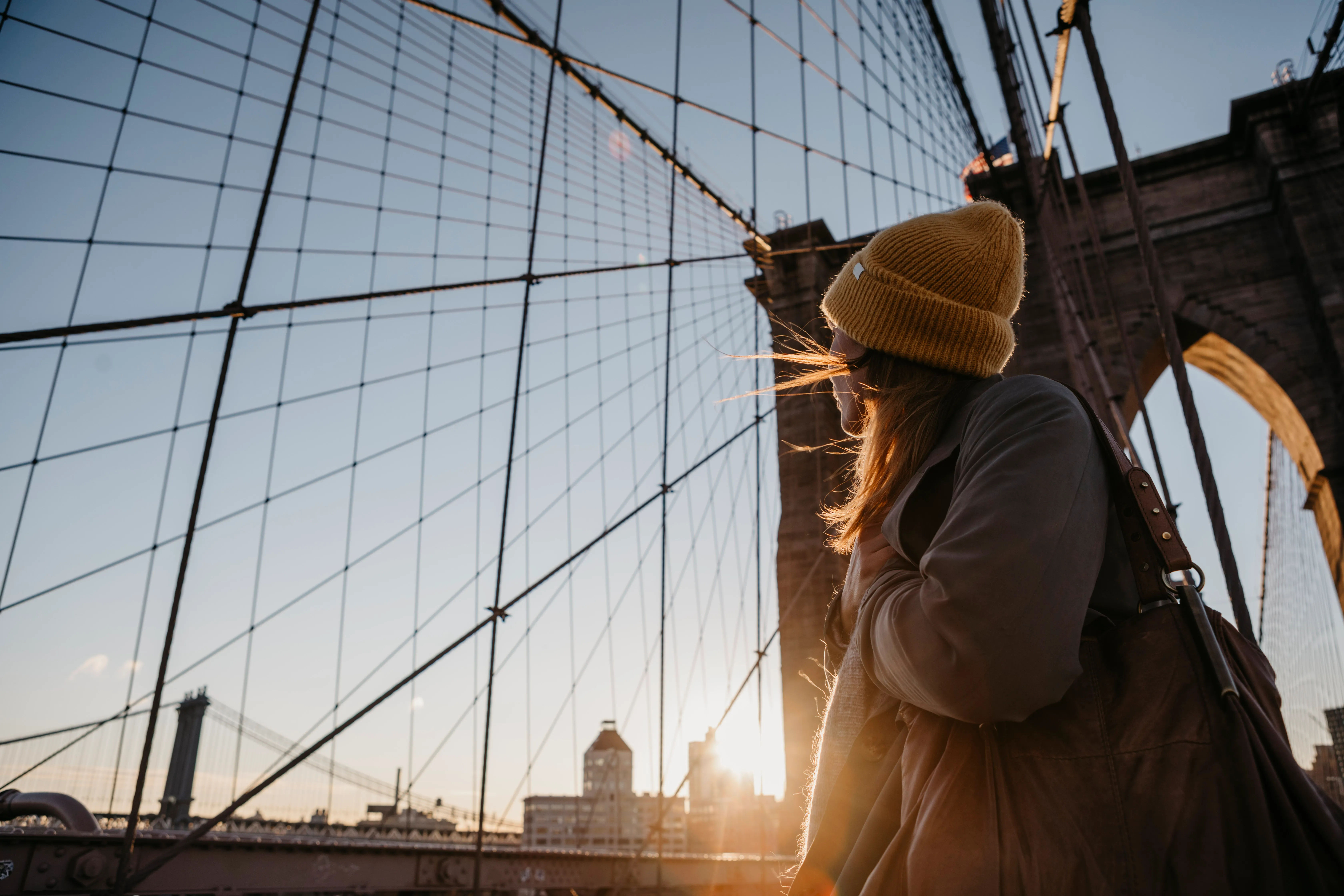 Woman in a knit hat walking across the Brooklyn Bridge at sunset
