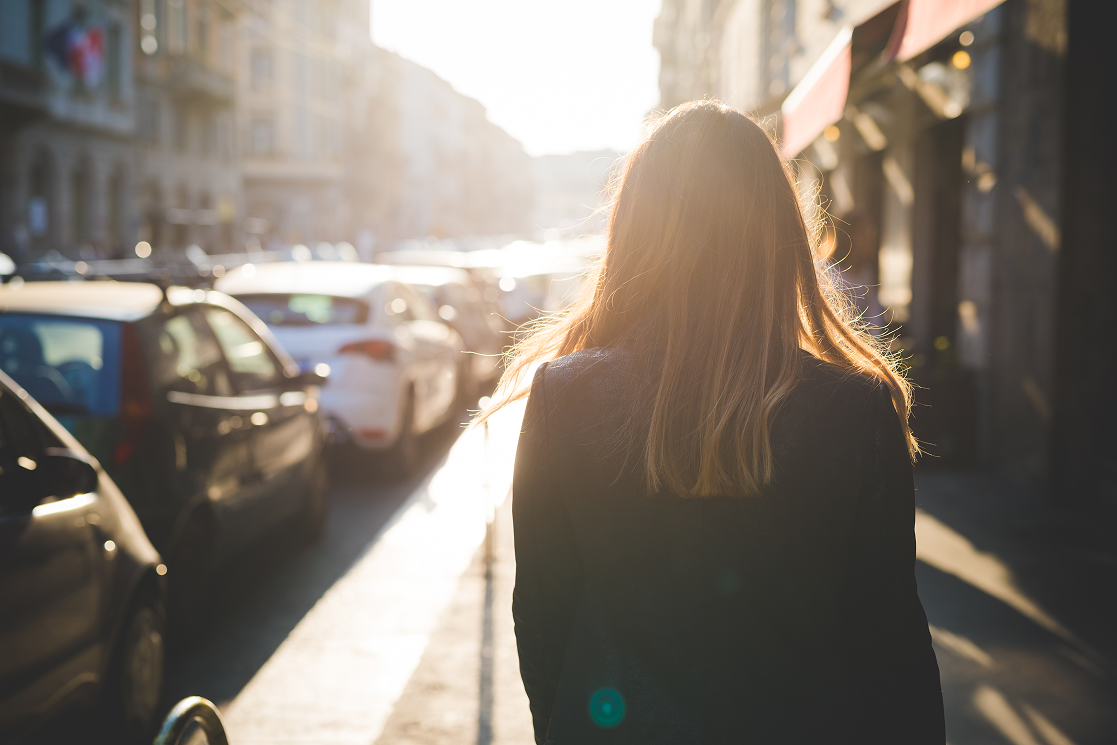 Woman walking down a sunlit city sidewalk lined with parked cars