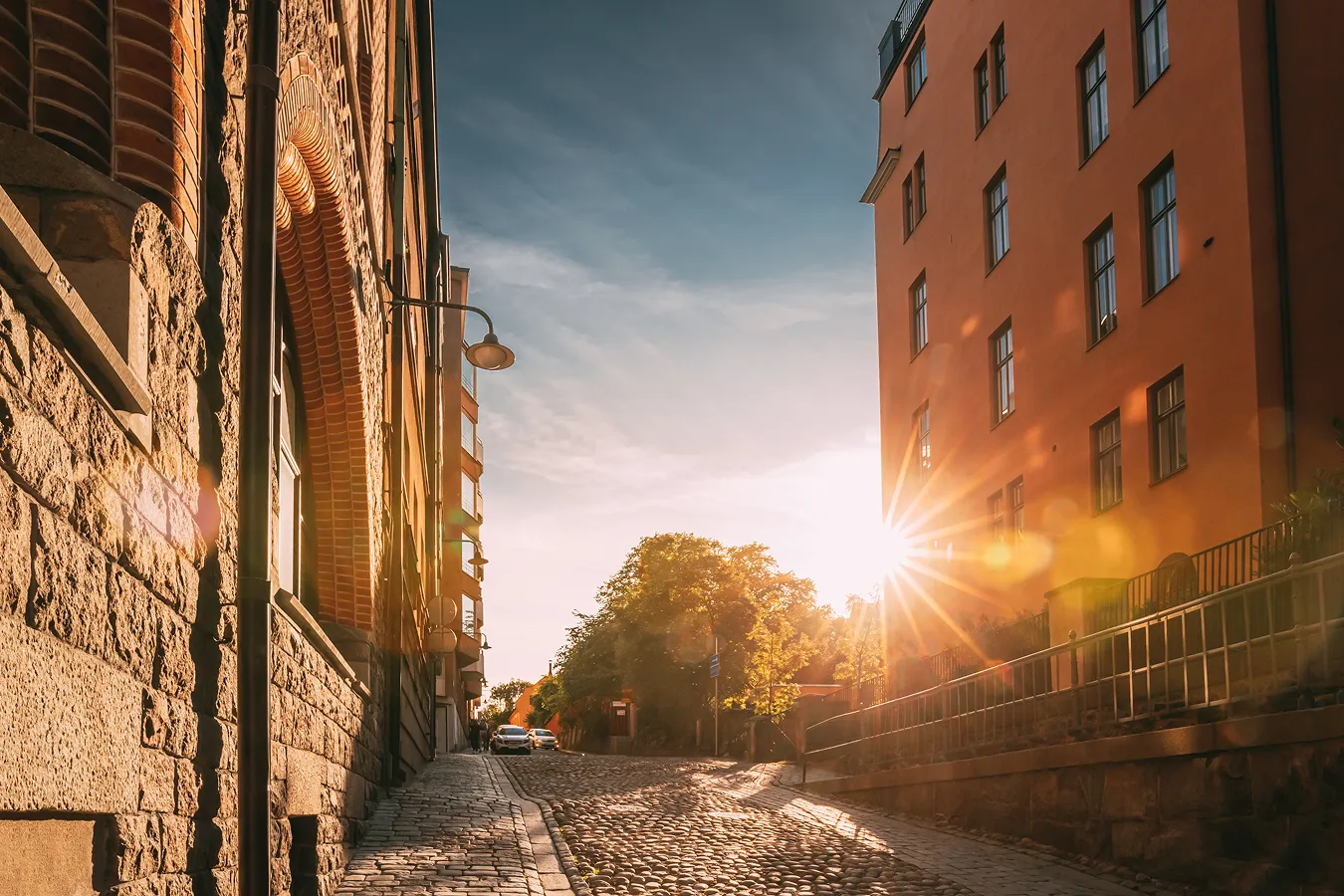 Cobblestone street between two buildings at sunset with the sun partially visible near a tall orange building and some trees in the background.