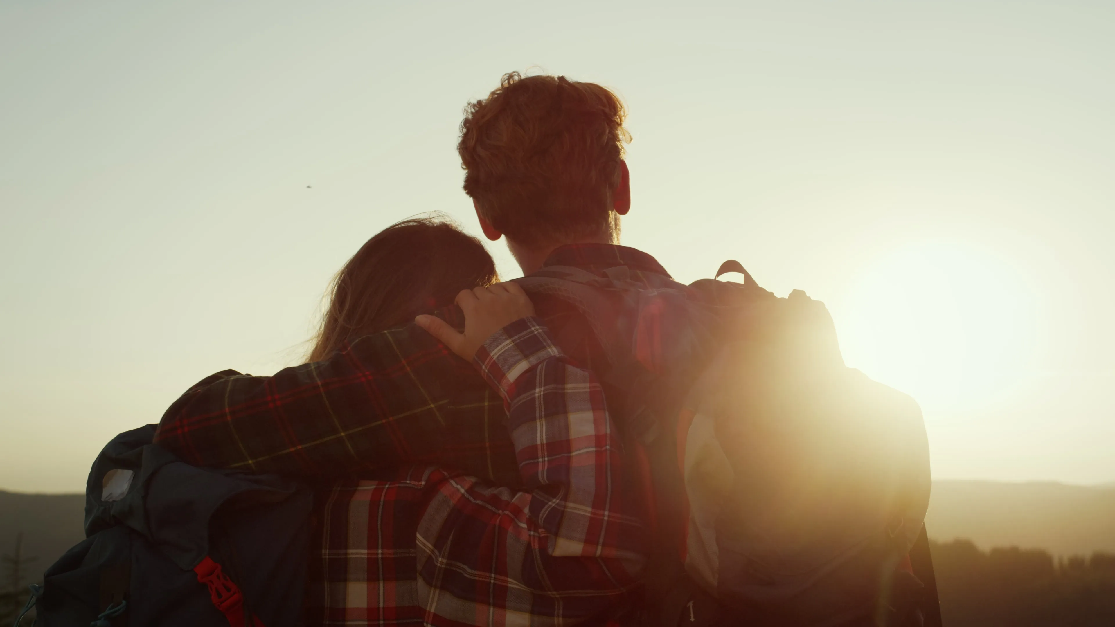 Back view of a couple with backpacks embracing as they watch a sunset over a landscape.