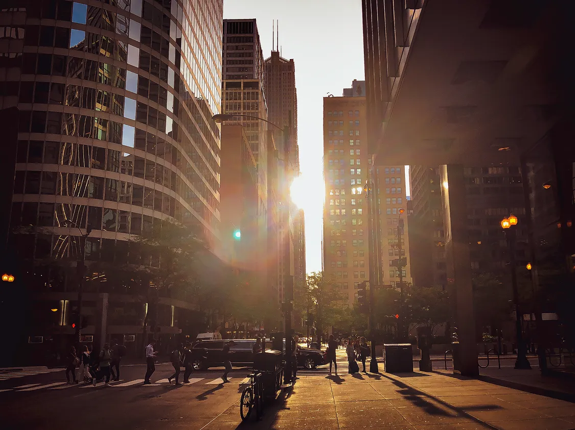 Sun setting between tall buildings in a city street with people crossing and shadows cast on the pavement.
