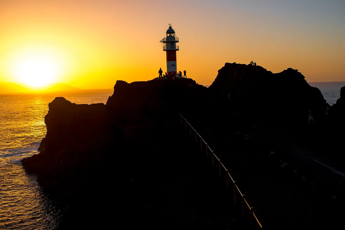 Silhouette of a lighthouse and rocky cliffs with people on top during a golden sunset over the ocean.