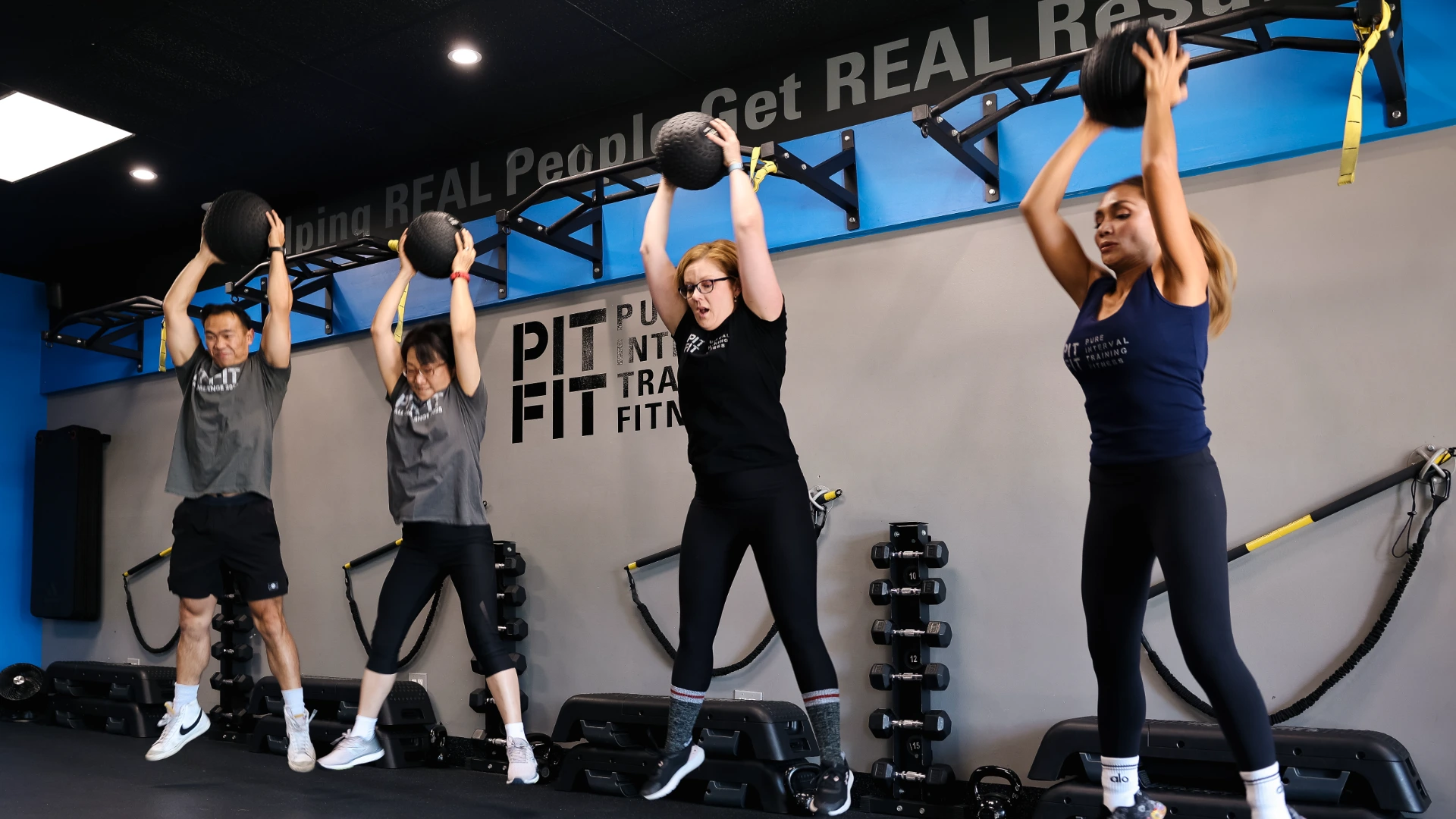 Four gym members performing simultaneous wall-ball overhead slams inside a fitness studio with blue and grey walls.