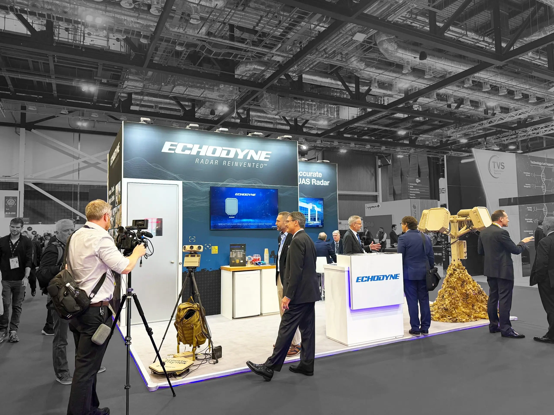 Trade show booth for Echodyne featuring radar display technology with a cameraman filming and several business professionals conversing.