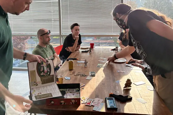 A group of adults gathered around a table playing a board game with cards and tokens in a sunlit room.
