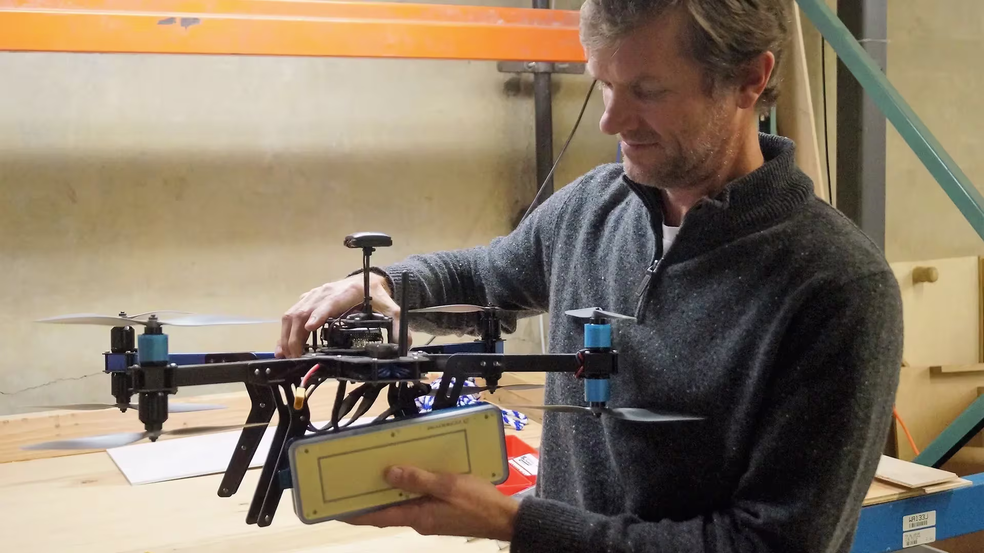 Man wearing dark sweater inspecting and holding a drone with four propellers in a workshop.