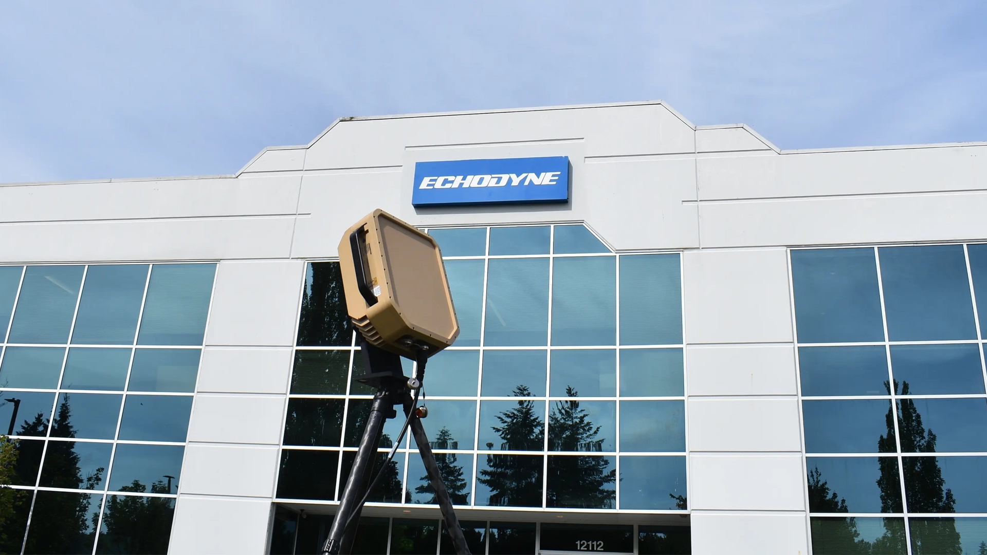 Modern white office building with large glass windows and a blue Echodyne sign above the entrance, with a radar device mounted on a tripod in front.