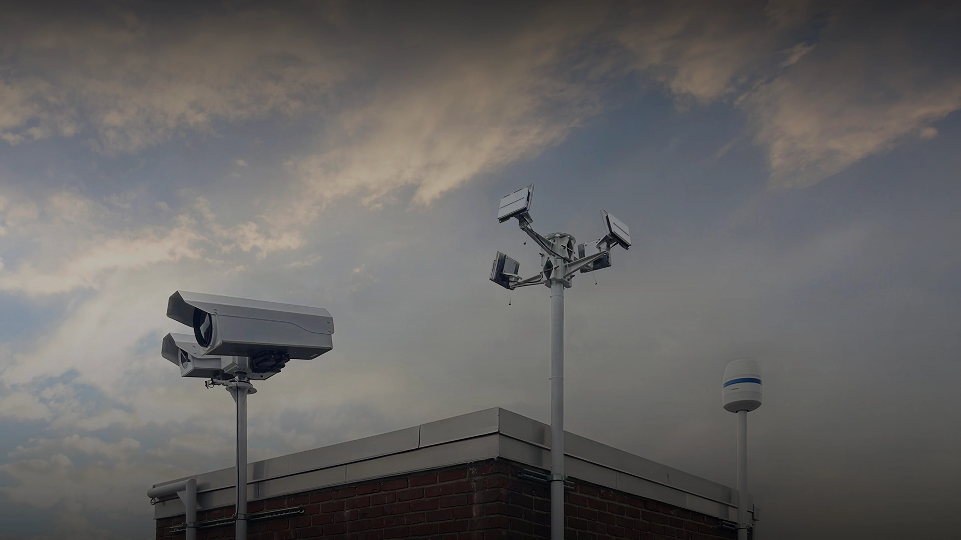 Security cameras and EchoGuard equipment mounted on poles at the corner of a brick building under a cloudy sky.