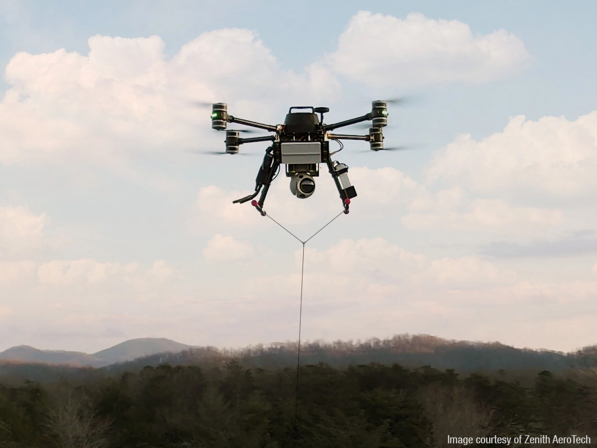 EchoFlight drone with a mounted camera flying over forested hills under a partly cloudy sky.