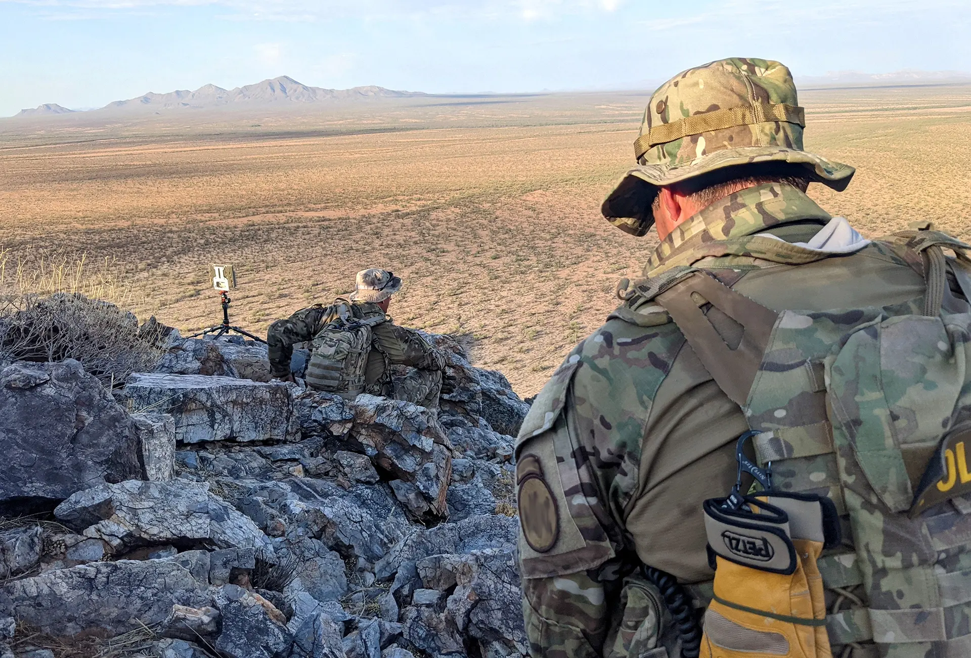 Two soldiers in camouflage gear positioned on rocky terrain overlooking a vast desert landscape with a small EchoGuard device on a tripod nearby.