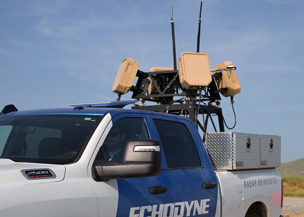 White and blue Echodyn radar truck with mounted EchoShield equipment on its roof under a clear sky.