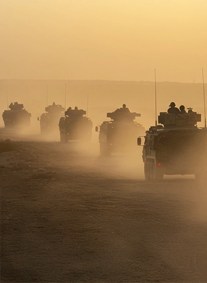A convoy of military armored vehicles moving through a dusty desert landscape at sunset.