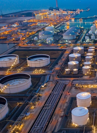 Aerial view of an industrial refinery complex with large storage tanks and pipelines lit at dusk.