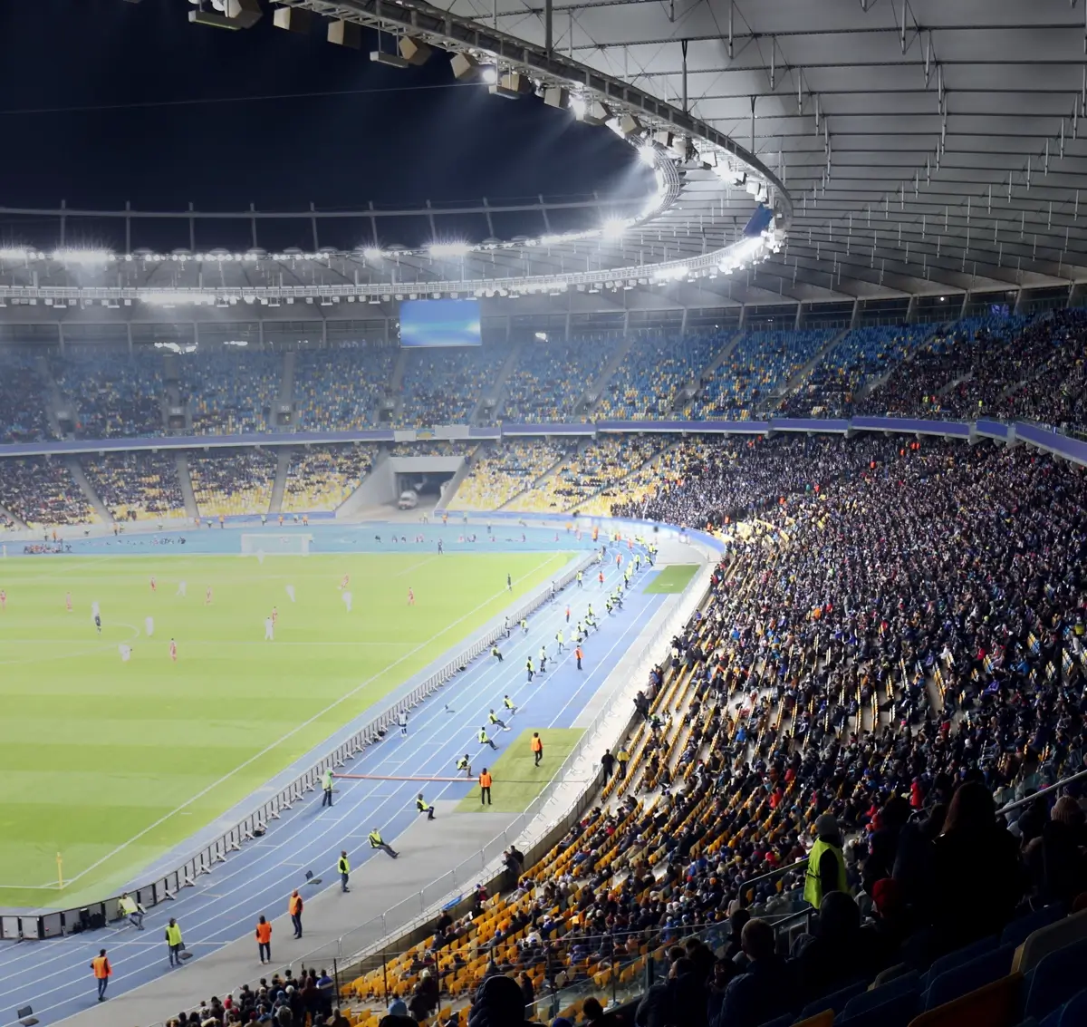 Nighttime view of a crowded stadium with a soccer match and bright overhead lights.