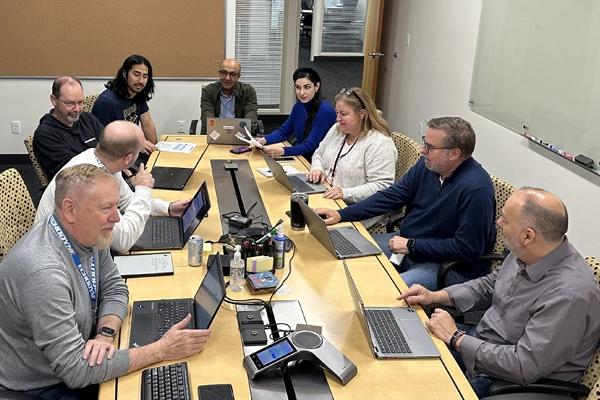 Eight people working and discussing around a conference table with laptops and documents.