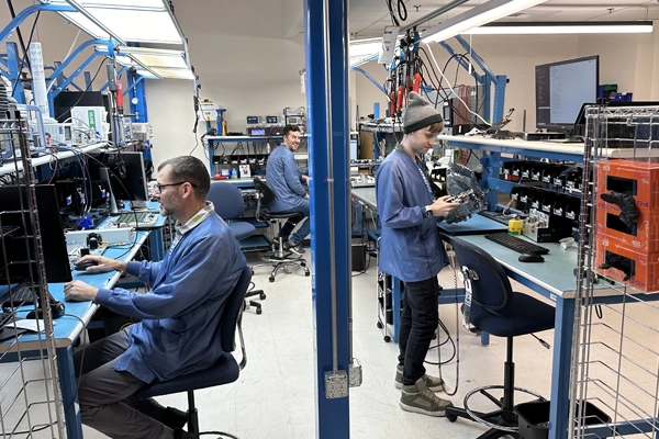 Three people in blue lab coats working with electronics in a tech lab with desks and monitors.